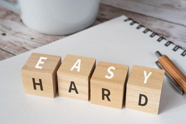 Wooden blocks with EASY and HARD text of concept, a pen, a notebook, and a cup.