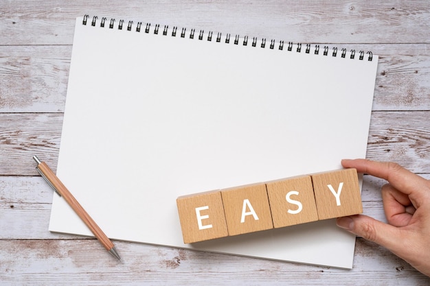 Wooden blocks with EASY text of concept, a pen, a notebook, and a hand.