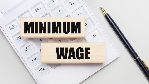 흰색 계산기의 밝은 배경에 MINIMUM WAGE Iie가 있는 나무 블록 Nearby는 검은색 손잡이 비즈니스 개념입니다.