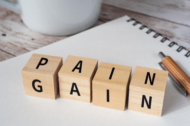 개념의 PAIN GAIN 텍스트, 펜, 노트북 및 컵이 있는 나무 블록.