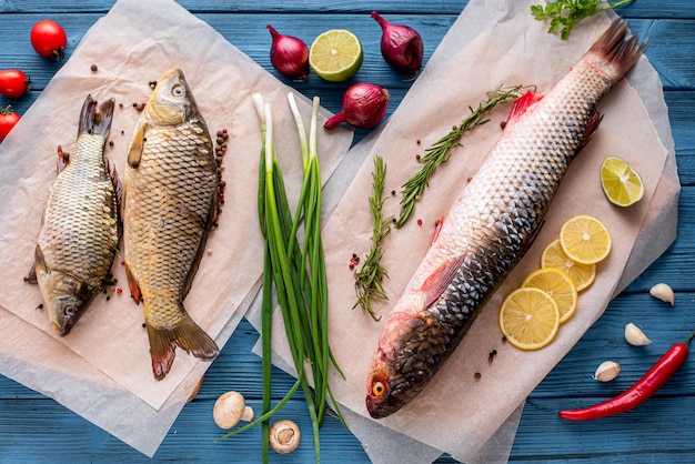 On a wooden blue background there is a fish pelengas sea fish and freshwater carp, next to spices fruits, vegetables and herbs