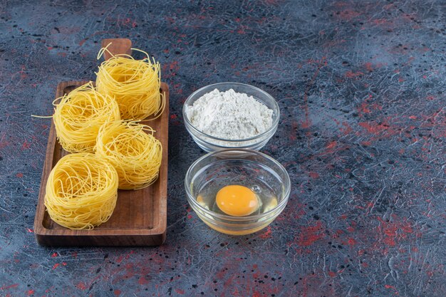 A wooden board of raw dry nest pasta with flour and uncooked egg on a dark surface .