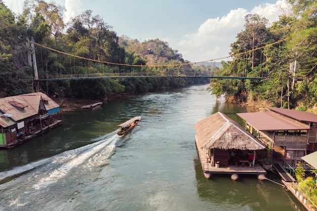 Wooden boat sailing on river kwai with tropical wooden village