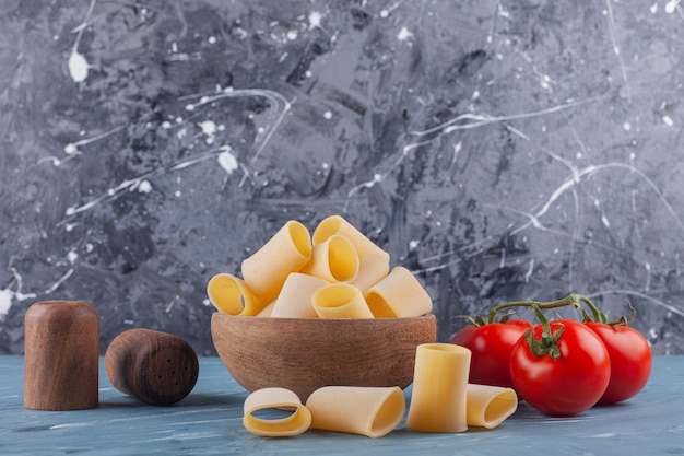 A wooden bowl of dry raw tube pasta with fresh red tomatoes and spices on a blue table .