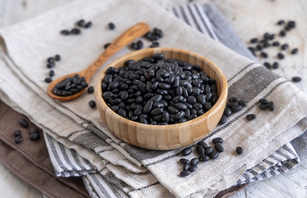 Wooden bowl full of dried black beans with a wooden spoon on kitchen towels on a wooden table close up. Healthy eating and vegetarian concept. Traditional Latin American cousin ingredient