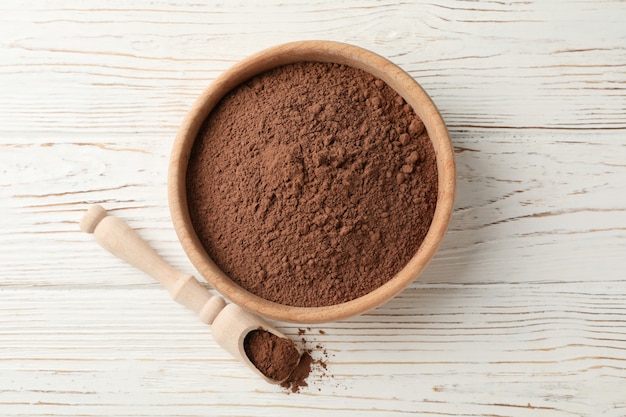 Wooden bowl and scoop with cocoa powder on white wood, top view