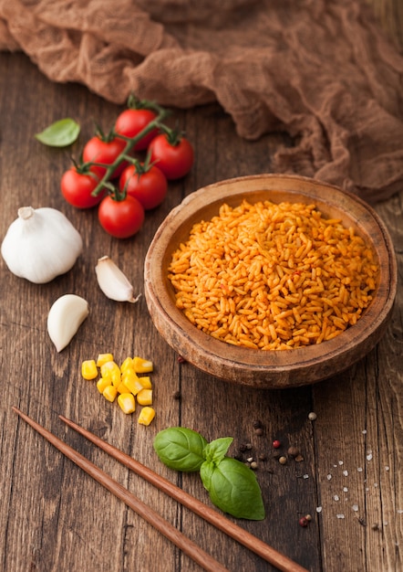 Wooden bowl with boiled red long grain basmati rice with vegetables on wooden table background with sticks and tomatoes with corn,garlic and basil.