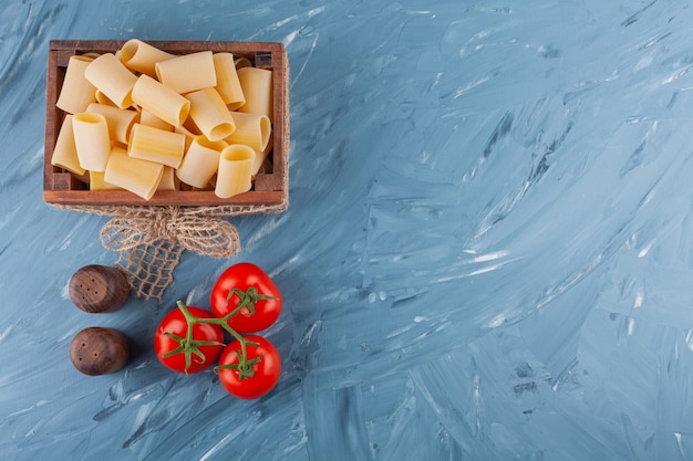 A wooden box of dry raw pasta with fresh red tomatoes on a marble table .