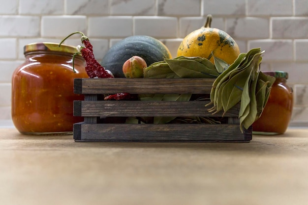 Wooden box with autumn vegetables and jars of pickles on the kitchen table Zucchini red hot peppers bay leaves and tomatoes Harvest season and preparations for the winter