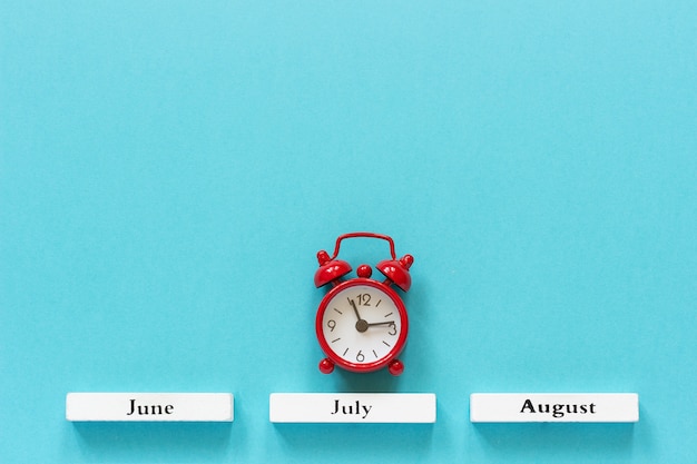 Wooden calendar summer months and red alarm clock over July on blue background. 