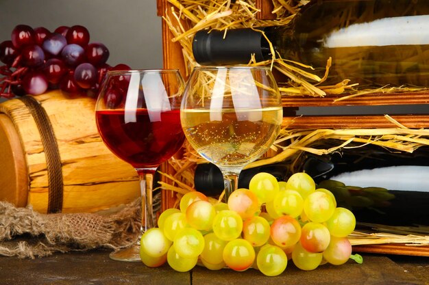 Деревянный ящик с винными бутылками, бочка, бокал и виноград на деревянном столе на сером фоне