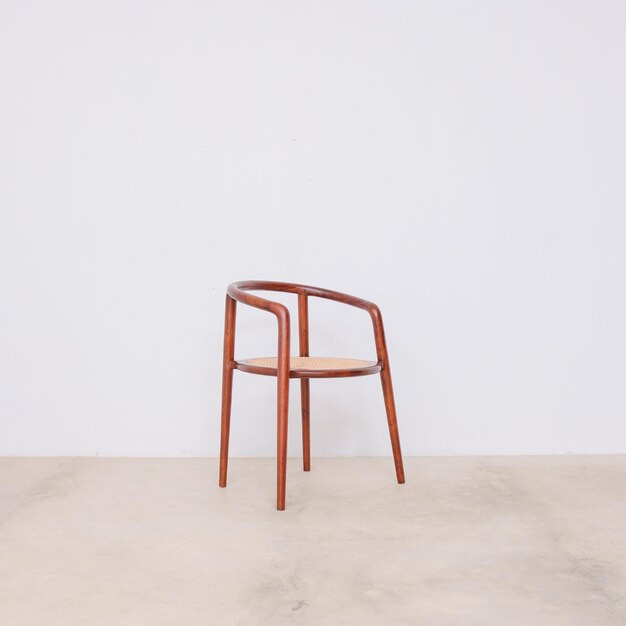 A wooden chair with a wooden seat sits in front of a white wall.