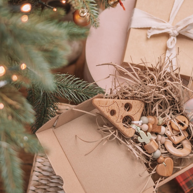 Wooden Christmas tree-shaped children's teether with silicone beads lies among gifts in open box under Christmas tree. Square frame instagram use