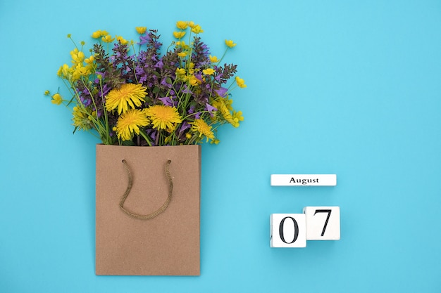 Wooden cubes calendar August 7 and field colorful rustic flowers in craft package on bluefor text and design