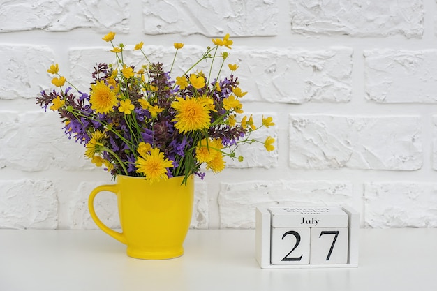 木製キューブカレンダー7月27日と白いレンガの壁に明るい色の花と黄色のカップ。テンプレートカレンダーの日付