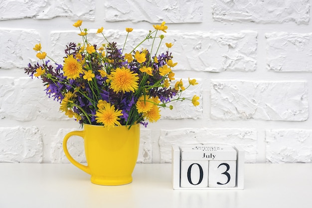 나무 큐브 달력 7 월 3 일 및 흰색 벽돌 벽에 밝은 색된 꽃과 노란색 컵.