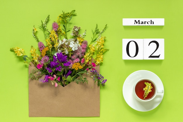 Photo wooden cubes calendar march 2. cup of herbs tea, kraft envelope with multi colored flowers on green background. concept hello spring creative top view flat lay