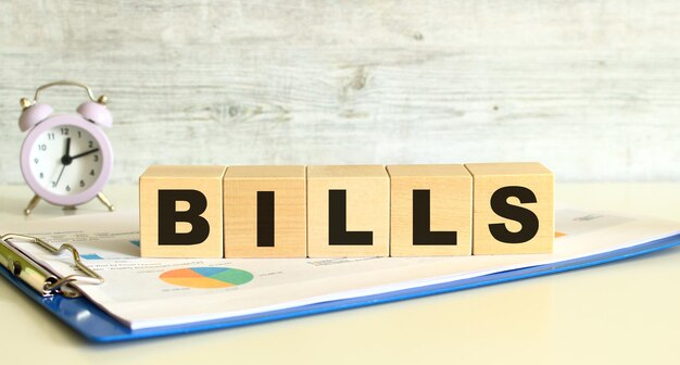 Wooden cubes lie on a folder with financial charts on a gray background The cubes make up the word BILLS Business concept