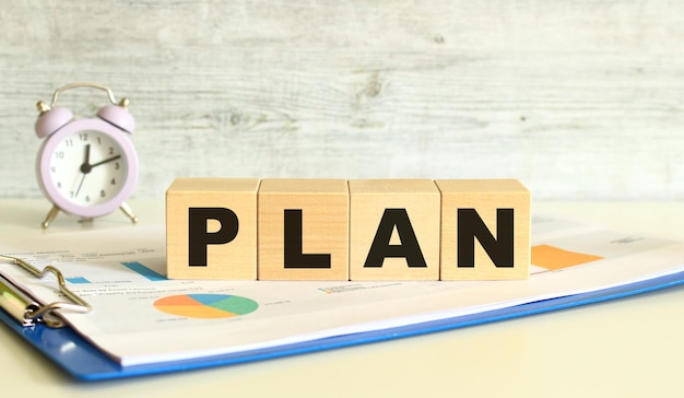 Wooden cubes lie on a folder with financial charts on a gray\
background the cubes make up the word plan