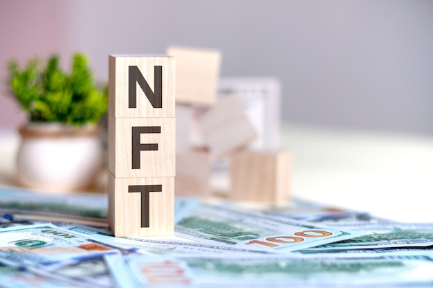 Wooden cubes with the letters NFT arranged in a vertical pyramid on banknotes, green potted plant. NFT - short for , business concept