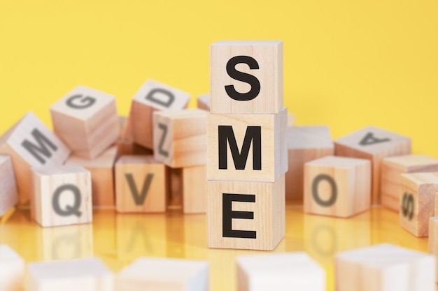 Cubi di legno con lettere sme disposti in una piramide verticale, sfondo giallo, riflesso dalla superficie del tavolo, concetto di business, sme - abbreviazione di piccole e medie imprese