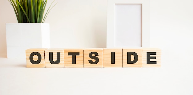 Wooden cubes with letters on a white table. The word is OUTSIDE. White background with photo frame, house plant.