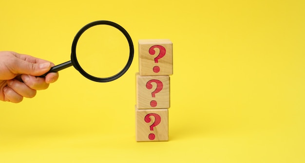 Wooden cubes with question marks and a female hand holds a magnifying glass on a yellow surface