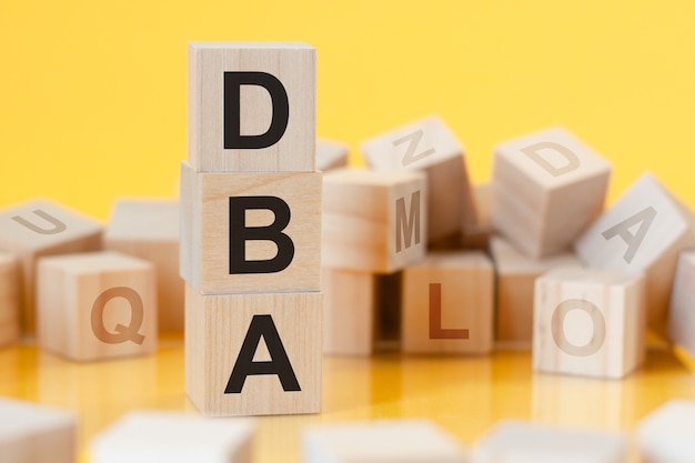 Фото Деревянные кубики со словом dba, расположенные в виде вертикальной пирамиды, желтая поверхность, отражение от поверхности стола
