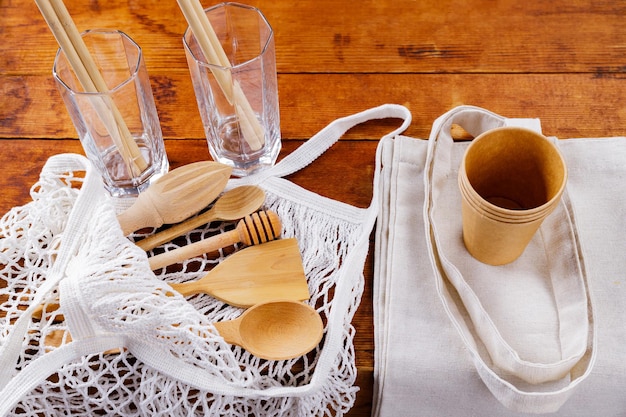 Wooden cutlery kitchenware and mesh bag on wooden table. Bamboo straws in empty glasses on a wooden boards. Disposable paper cups and linen shopping bag. Zero waste concept