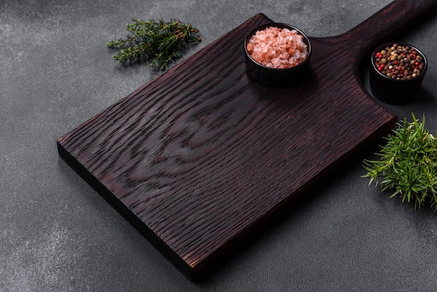 Wooden cutting board salt pepper spices and herbs on a dark concrete background