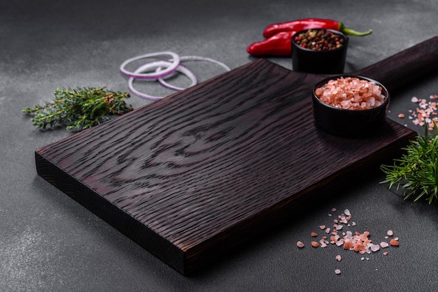 Photo wooden cutting board salt pepper spices and herbs on a dark concrete background