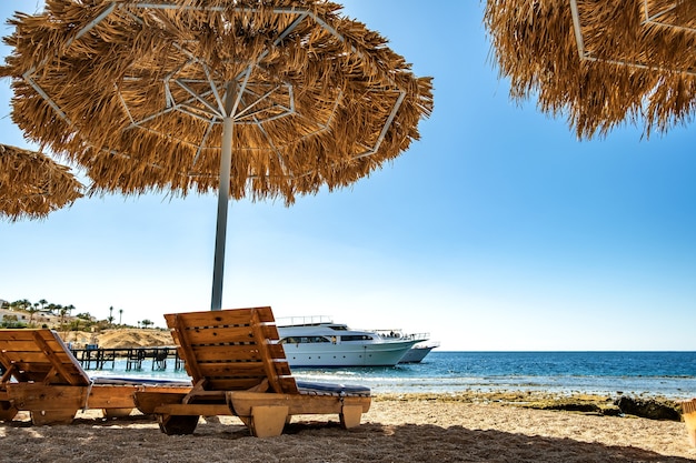 Wooden deck chairs under rough straw sun umbrella on sea beach and big white yacht ship in water near shore on sunny summer day.