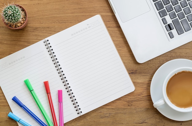 Wooden desk table with notebook, computer laptop, color pens, cactus and cup of coffee. Top view with copy space.