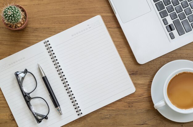 Wooden desk table with notebook, computer laptop, pen, eyeglasses, cactus and cup of coffee. Top view with copy space