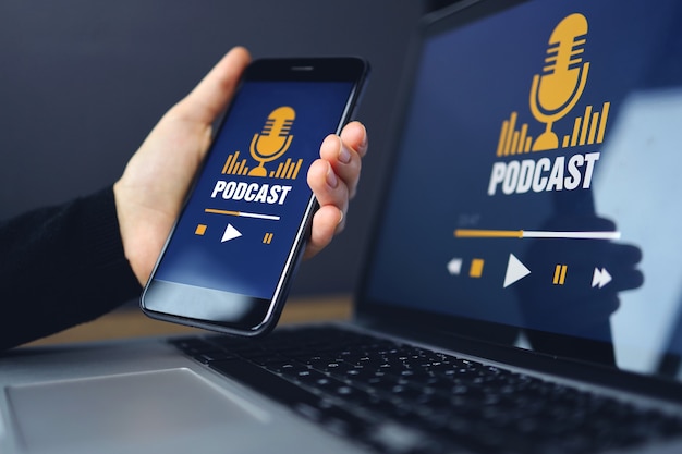 Wooden desk with a laptop computer with podcast website service in the screen. Hand holding smartphone with podcast app.