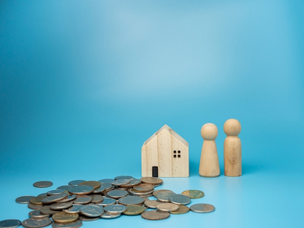 A wooden doll standing next to a replica wooden house and a pile of money on blue