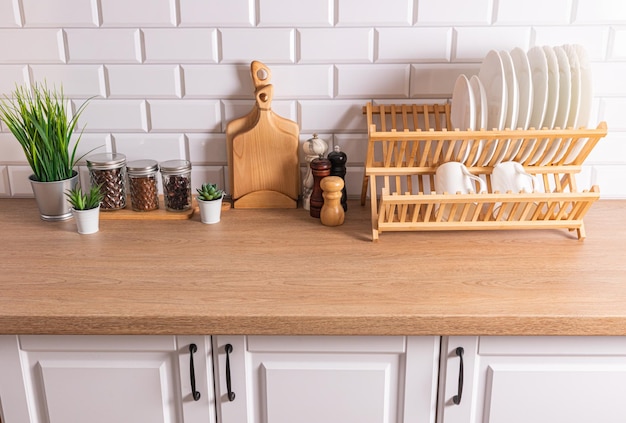 A wooden dryer with plates glass jars with bulk products spice mills on a wooden countertop opposite a white brick wall Eco items