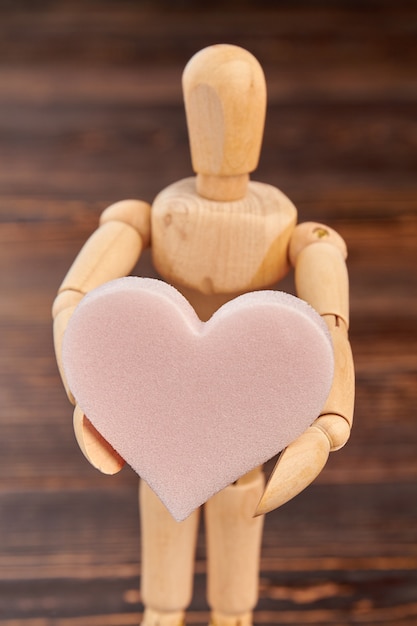 Wooden dummy holding pink soft heart. Wooden person standing with heart shaped sponge. Valentines holiday concept.