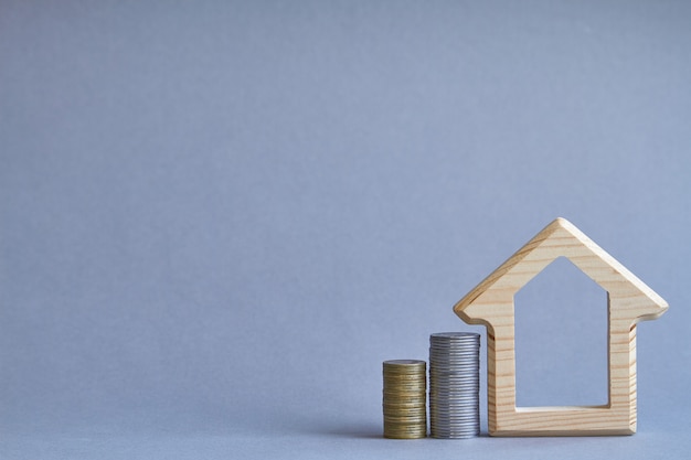 A wooden figurine of house with two columns of coins nearby on gray