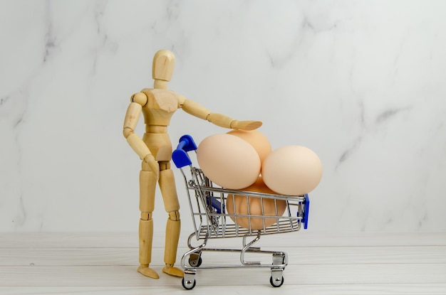 Wooden figurine of a man with a grocery cart filled with chicken eggs. Wooden mannequin with a basket of eggs. Easter preparation concept, grocery shopping, farming support. Space for text