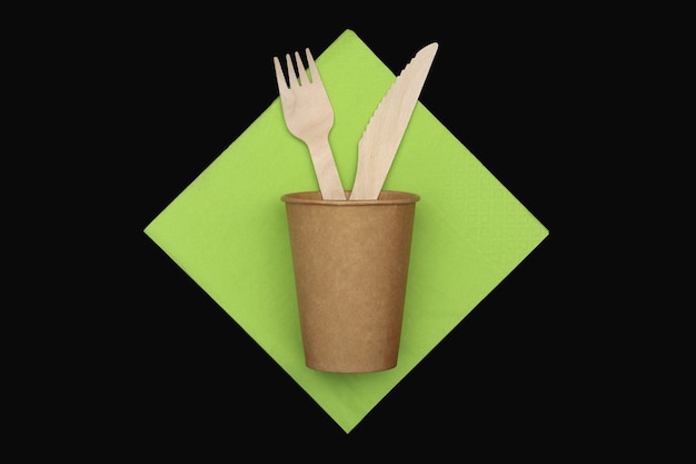 Photo wooden fork and knife in paper cup on green napkin on black background, top view.