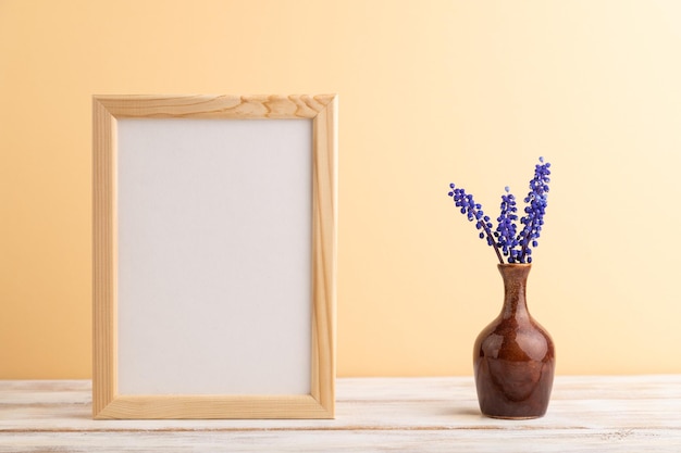 Wooden frame with blue muscari hyacinth flowers in ceramic vase\
on orange pastel background side view copy space mockup