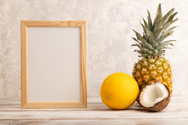 Wooden frame with pineapple, melon, coconut on gray concrete background. Side view, copy space. 