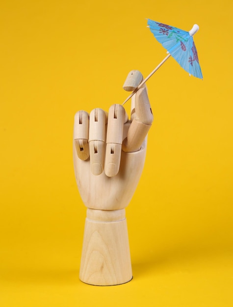 Wooden hand holding cocktail umbrella on yellow background