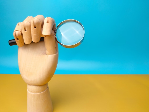 Wooden hand holding magnifying glass on yellow and blue background