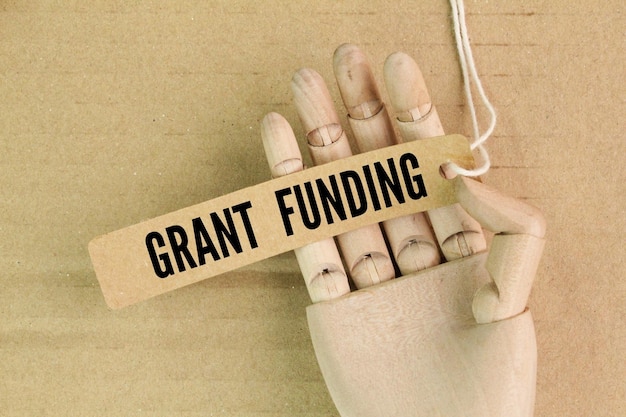Wooden hand and paper tag with the word GRANT FUNDING any funding or other aid or assistance from an