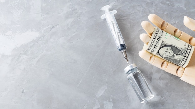 Wooden hand with vaccine bottle, syringe and dollar bill on gray background