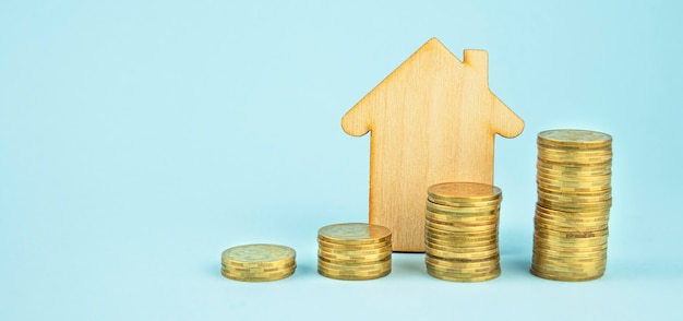 Wooden house and coins stacks on light blue background