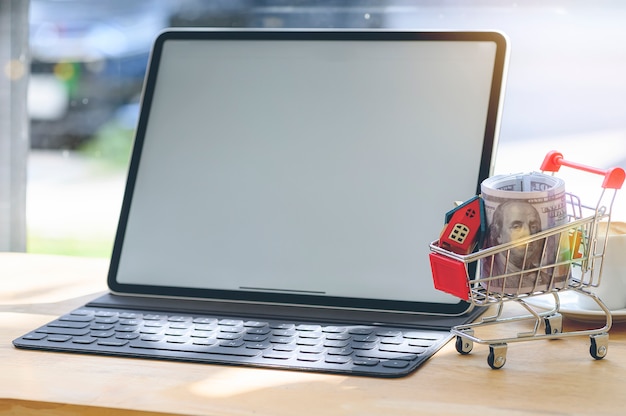 Wooden house model and bank note in shopping cart on wooden table with blank screen table.