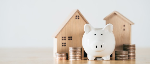 Wooden house with piggy bank and stacking coins on wooden table saving money for buying house